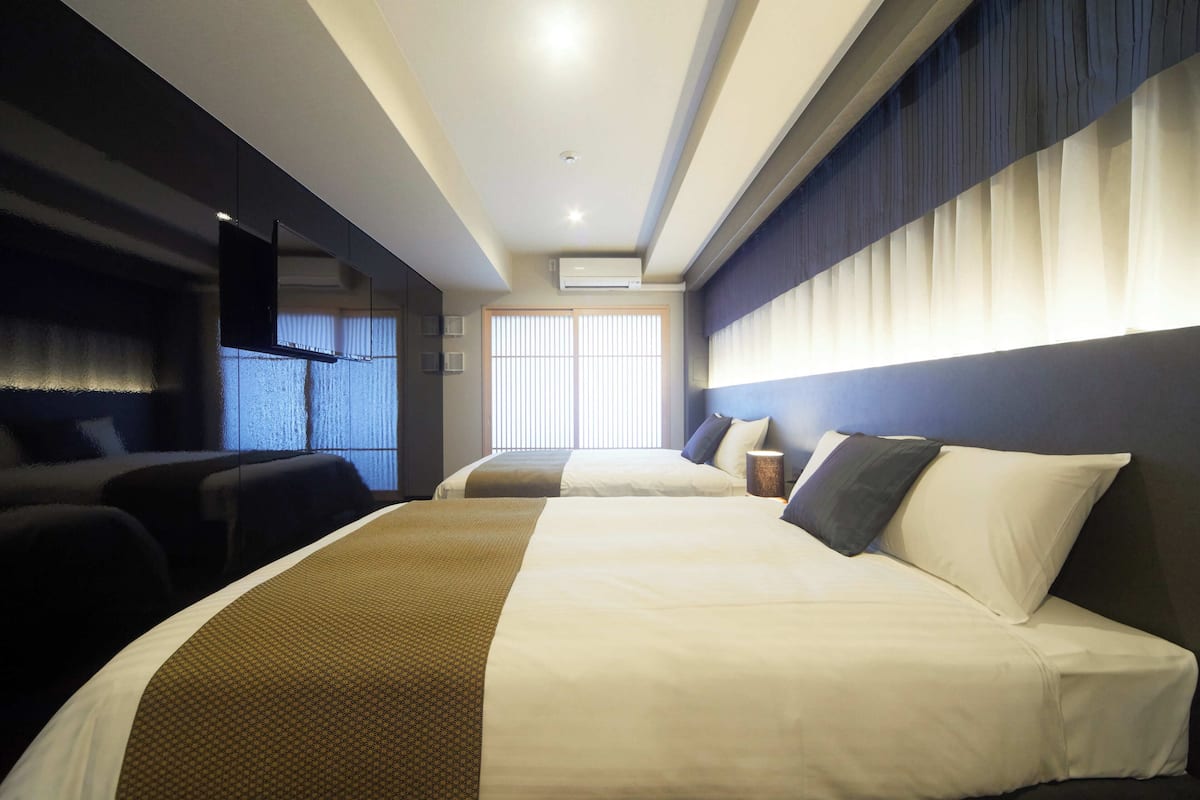 A hotel room with two beds, a flat-screen TV, a window with blinds, and a wall-mounted air conditioner.