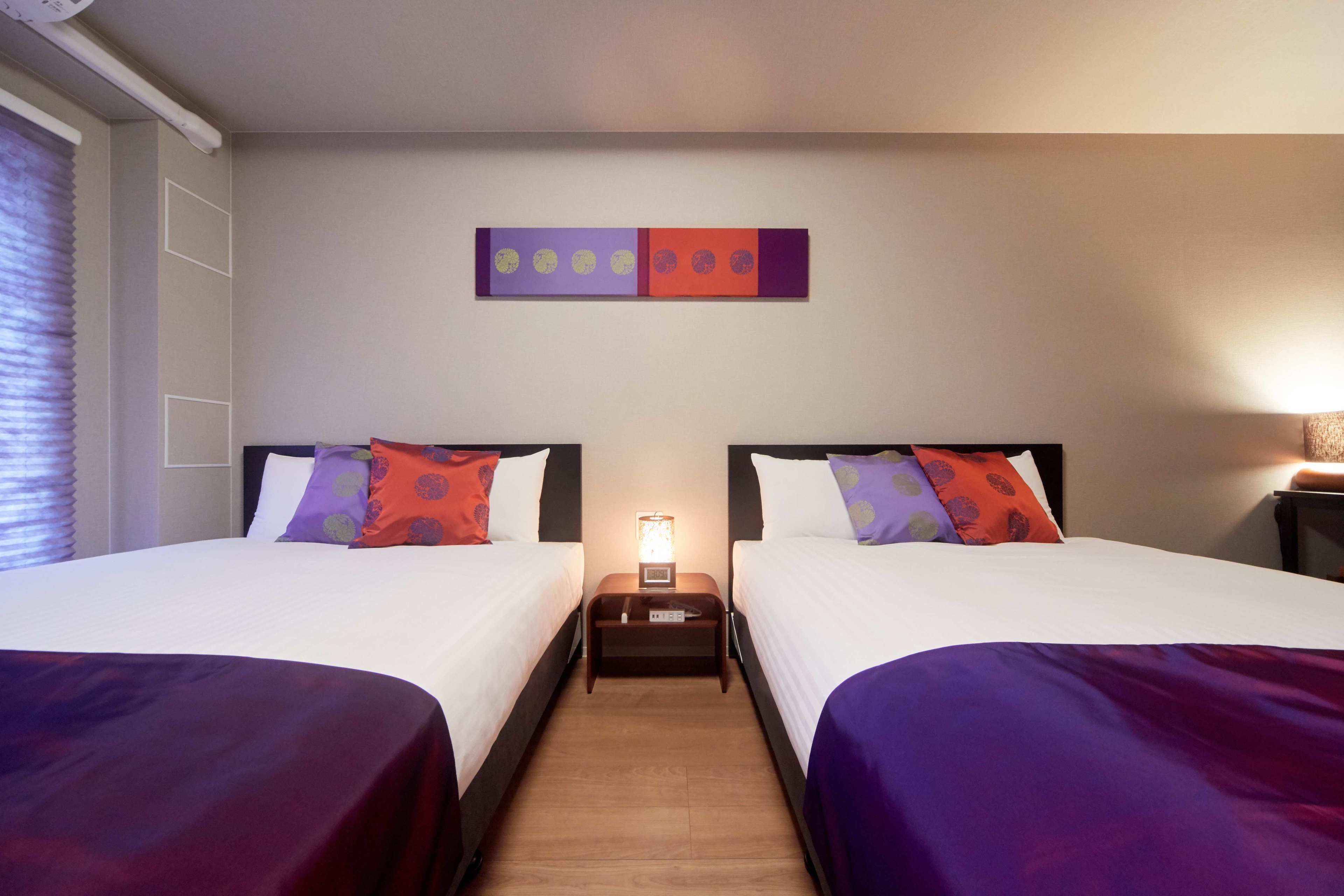 A hotel room with two beds, each with a purple bedspread, white linens, and multiple pillows. There is a nightstand between the beds with a lamp and a small table.