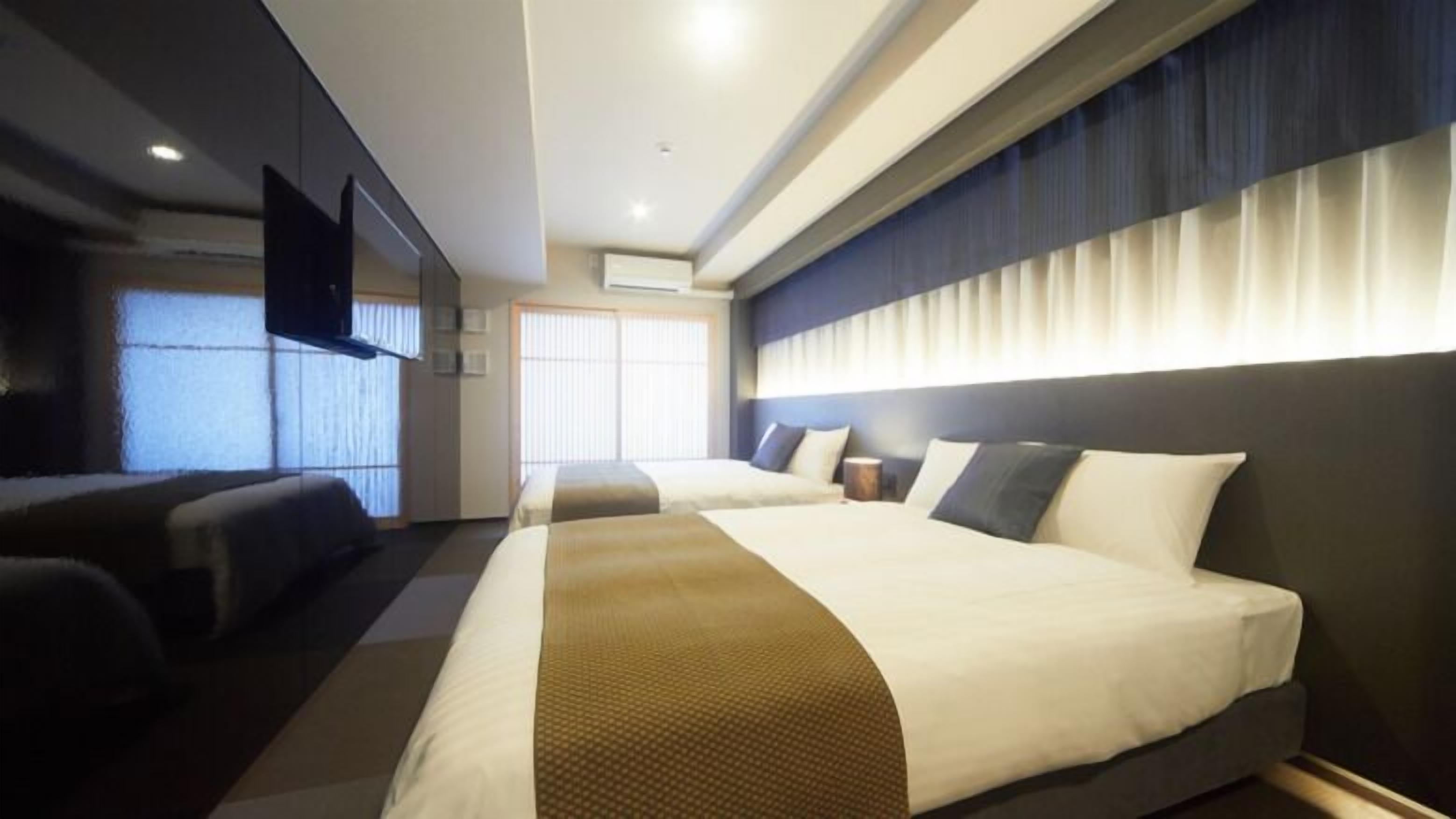 A hotel room with two beds, a flat-screen TV, a large window with blinds, and a ceiling-mounted air conditioning unit.