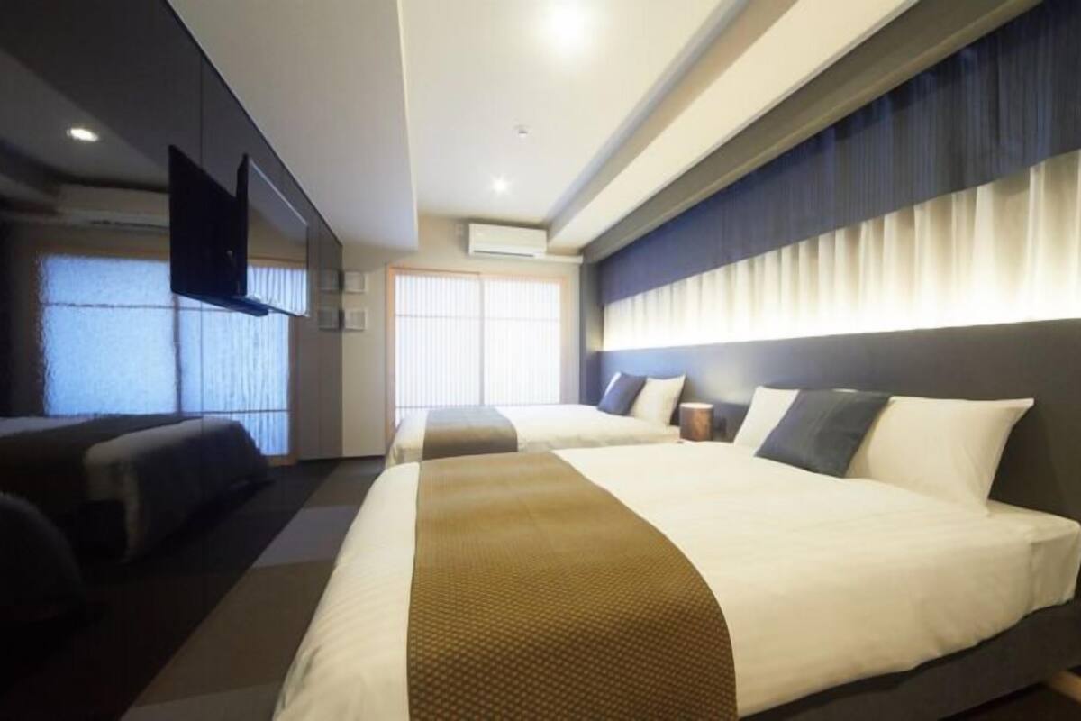 A hotel room with two beds, a flat-screen TV, a large window with blinds, and a ceiling-mounted air conditioning unit.