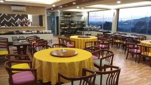 A dining area with round tables covered in yellow tablecloths, wooden chairs with cushioned seats, and a large window offering a view of the cityscape. - Nite & Day Laguna Bintan - Tanjungpinang (Tanjung Pinang)