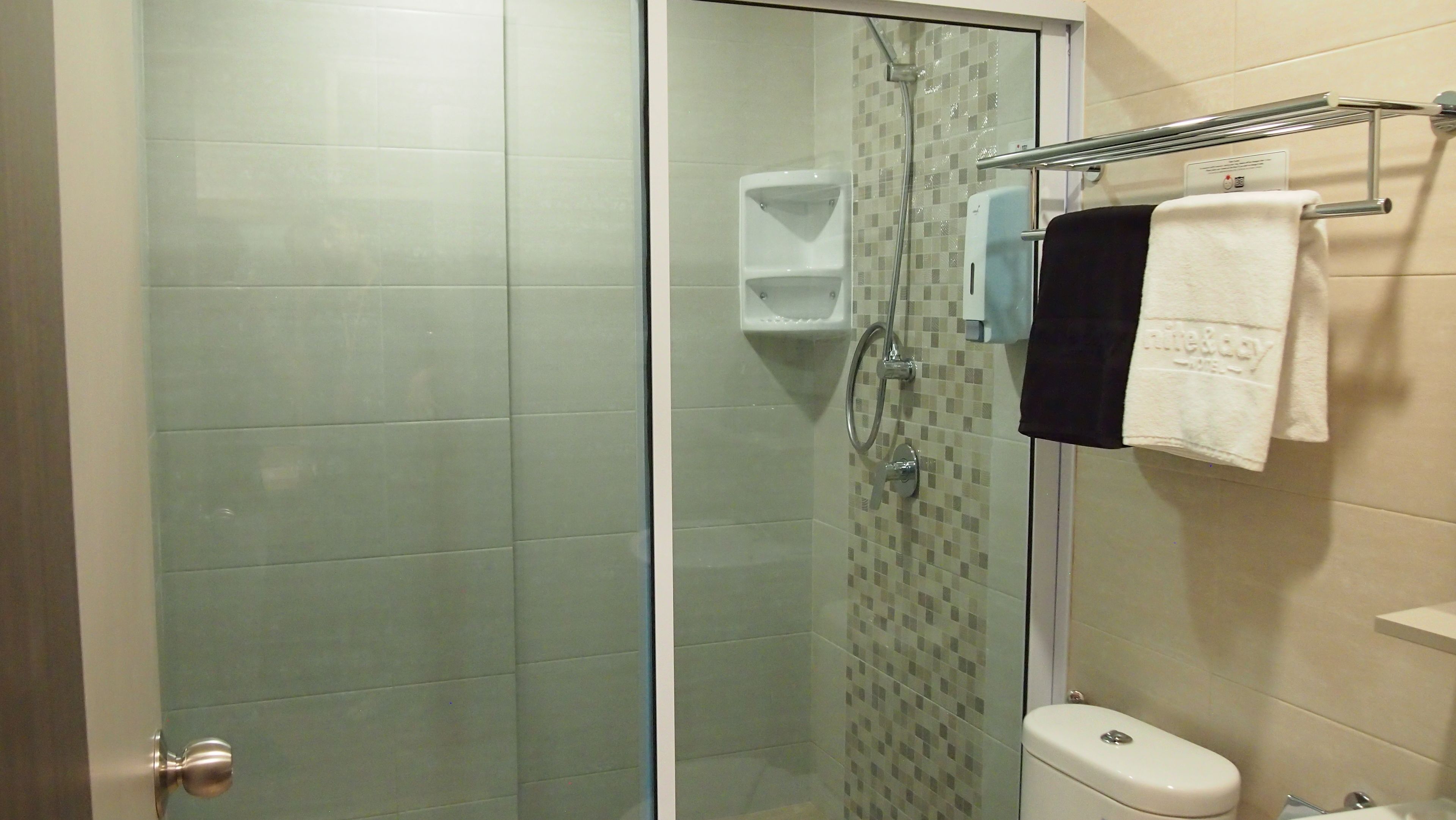 Un baño con cabina de ducha de vidrio, inodoro, toallero con dos toallas y dispensador de jabón.