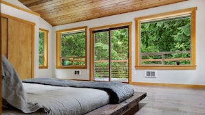 A bedroom with a large bed, wooden ceiling, and large windows with a view of greenery.