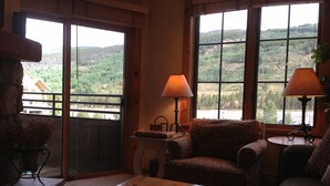 Ein gemütliches Wohnzimmer mit einem Holztisch, zwei gemusterten Sesseln und einem Sofa. Große Fenster bieten Blick auf eine bewaldete Landschaft.