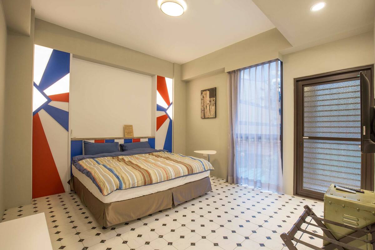 A bedroom with a bed, a suitcase, a window with curtains, a painting on the wall, and a geometric-patterned floor.