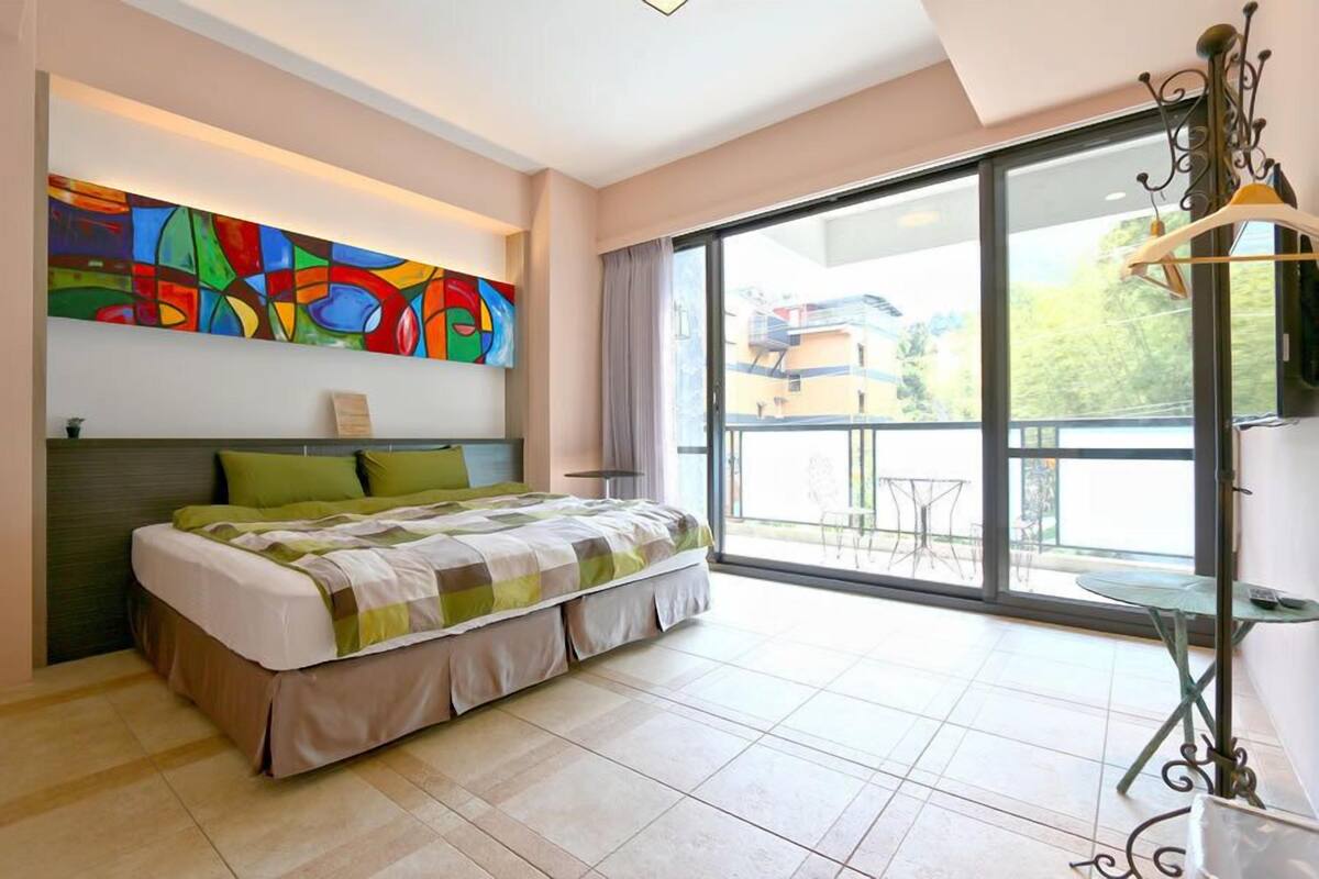 A bedroom with a large bed, a colorful abstract painting, a balcony with a table, and a view of buildings outside.