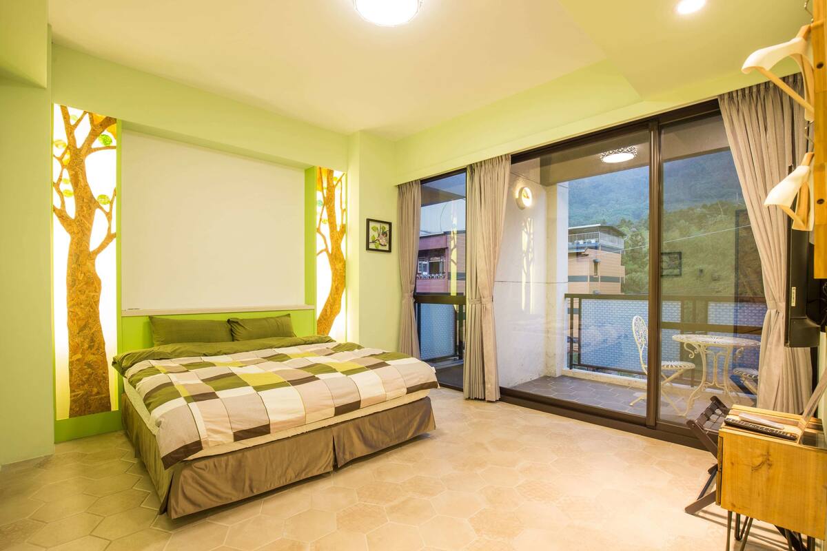 A bedroom with a bed, a window with curtains, a view of the outside, and a small table with a lamp.