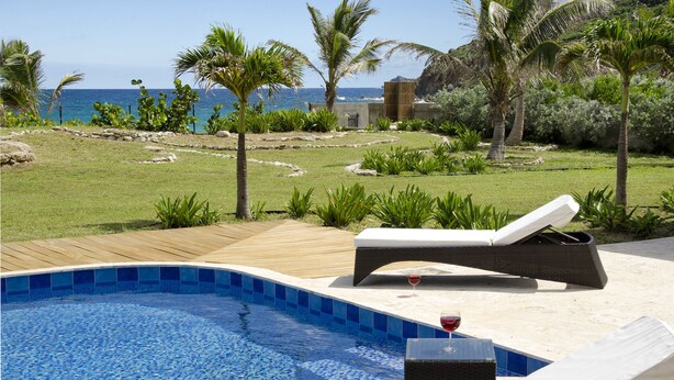 Área junto à piscina com espreguiçadeiras e um copo de vinho tinto, com vista para uma praia e oceano.