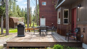 A wooden deck with a barbecue grill, a table with chairs, and a red building with a door and windows. - Shepherd Family Retreat- Fox | Main Home & Guest House sleeps 12 Near Yellowstone (West Yellowstone)