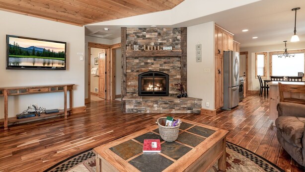 A spacious living room with a stone fireplace, wooden flooring, and a television mounted on the wall. - Shepherd Family Retreat- Fox | Main Home & Guest House sleeps 12 Near Yellowstone (West Yellowstone)