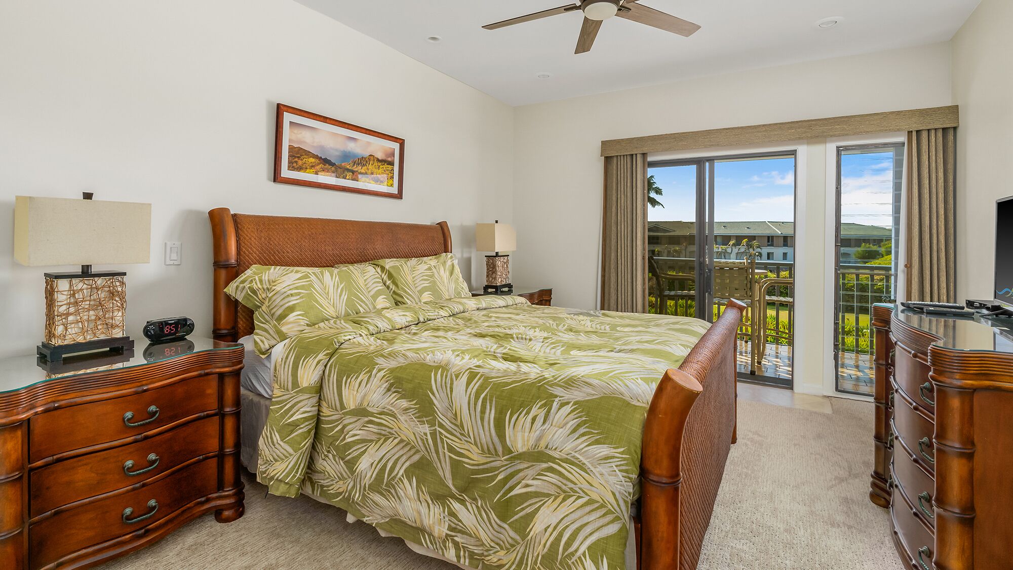 A bedroom with a bed, wooden nightstands, a dresser, a ceiling fan, a painting, and a balcony with a view.