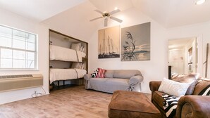 Ein gemütliches Wohnzimmer mit Deckenventilator, einem großen Fenster, einer Couch, einem braunen Sessel und einem Etagenbett mit weißem Kopfteil.
