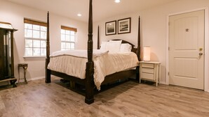 Ein Bett mit Himmel, dessen Gestell aus Holz gefertigt ist und das mit weißen Leinen bezogen ist, ein Nachttisch mit einer Lampe sowie eine Tür mit Schloss.