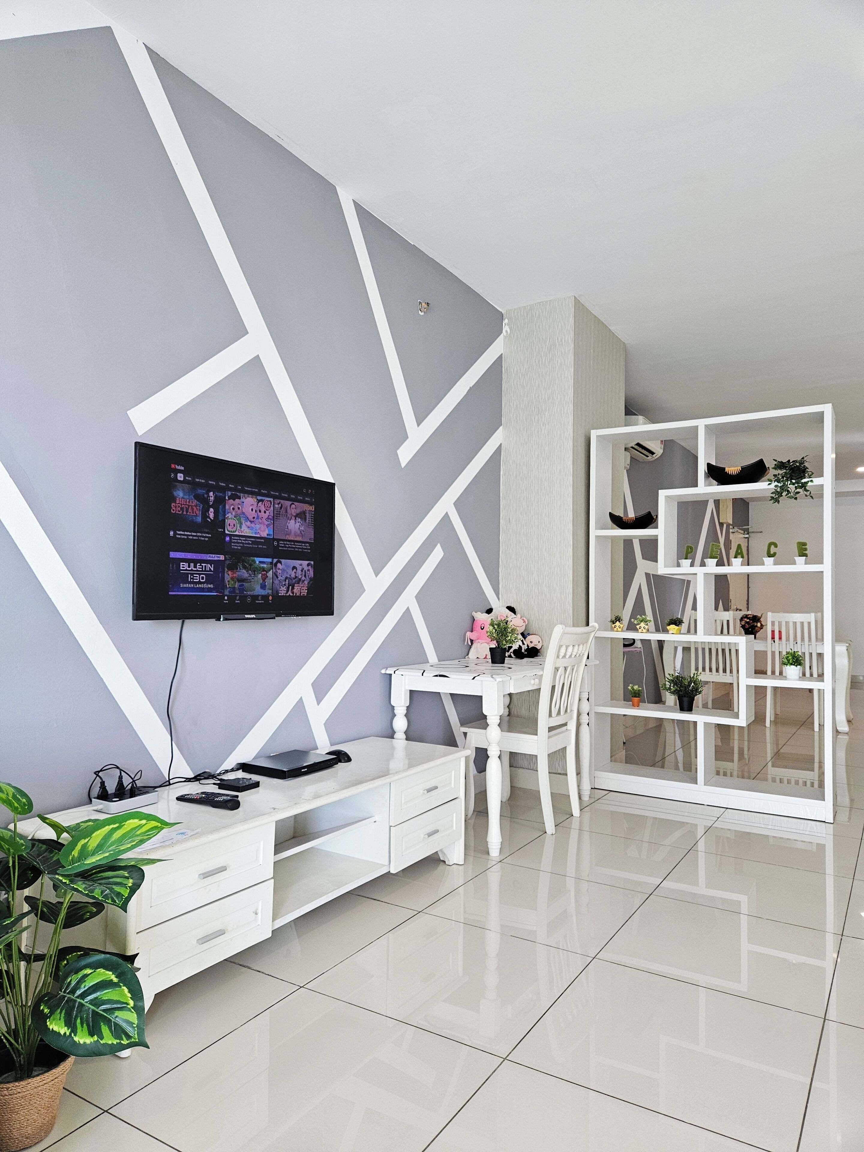A modern living room with a flat-screen TV mounted on the wall, a white desk with a chair, a white shelving unit with decorative items, and a potted plant.