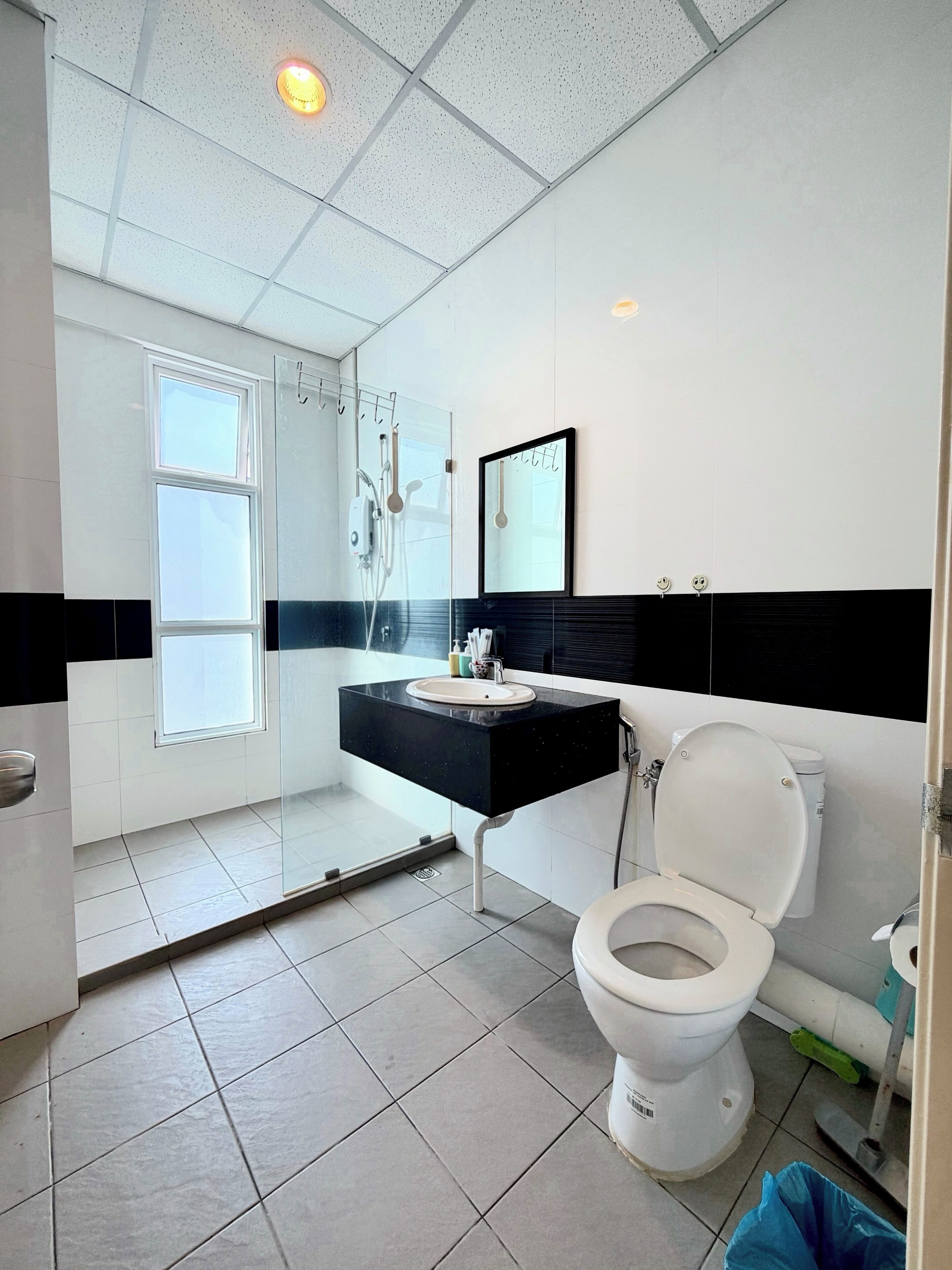 A modern bathroom with a black countertop, white sink, and a toilet. There is a shower area with a glass door and a window above it.
