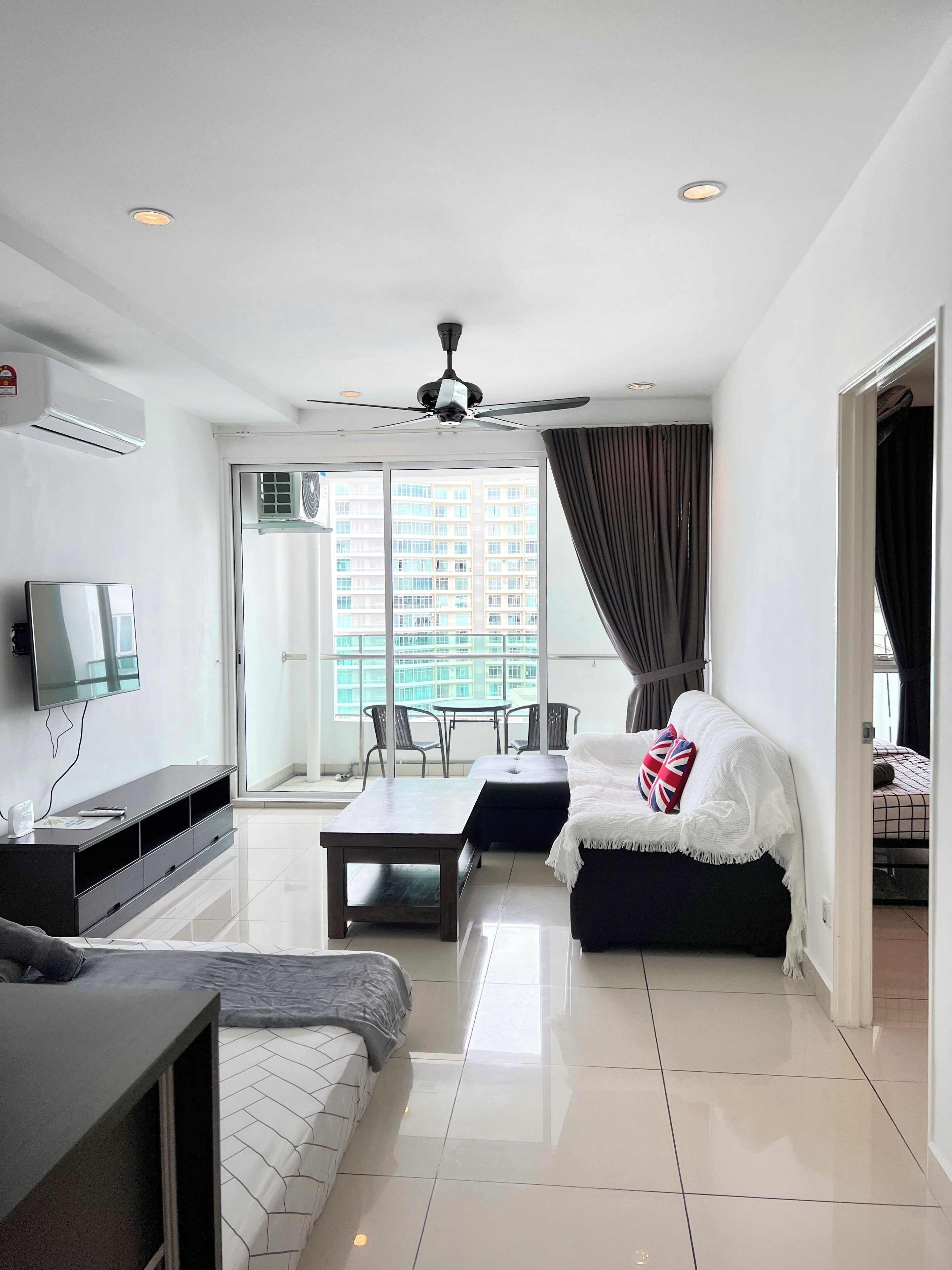 Una habitación de hotel moderna con televisión de pantalla plana, sofá, cama con almohada con la bandera Union Jack, mesa de comedor y balcón con vistas a un edificio de gran altura.