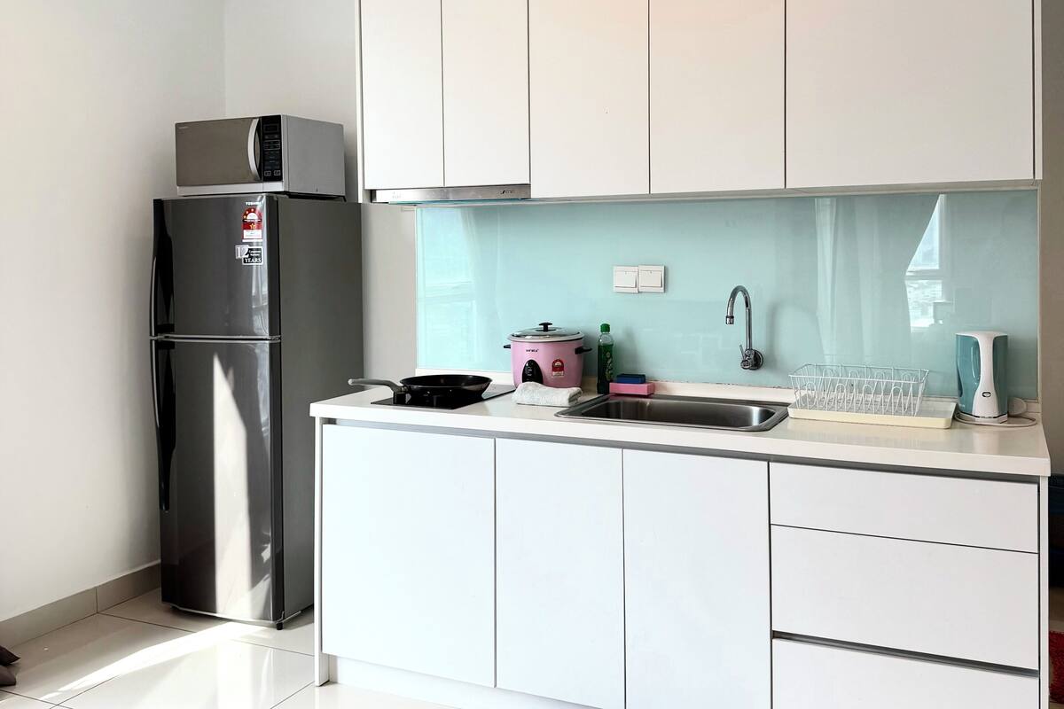 Eine moderne Küche mit weißen Schränken, einem Edelstahl-Kühlschrank, einem rosa Schongerät, einer schwarzen Bratpfanne, einem Spülbecken und einem Geschirrständer.