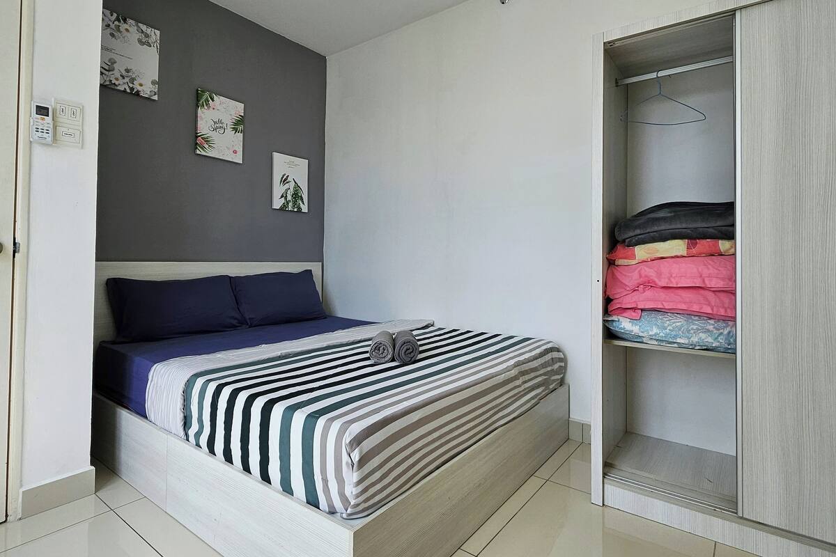 Ein ordentlich bezogenes Bett mit einem gestreiften Bettüberwurf, eine graue Wand mit zwei gerahmten Bildern und ein Kleiderschrank mit gefalteter Kleidung.