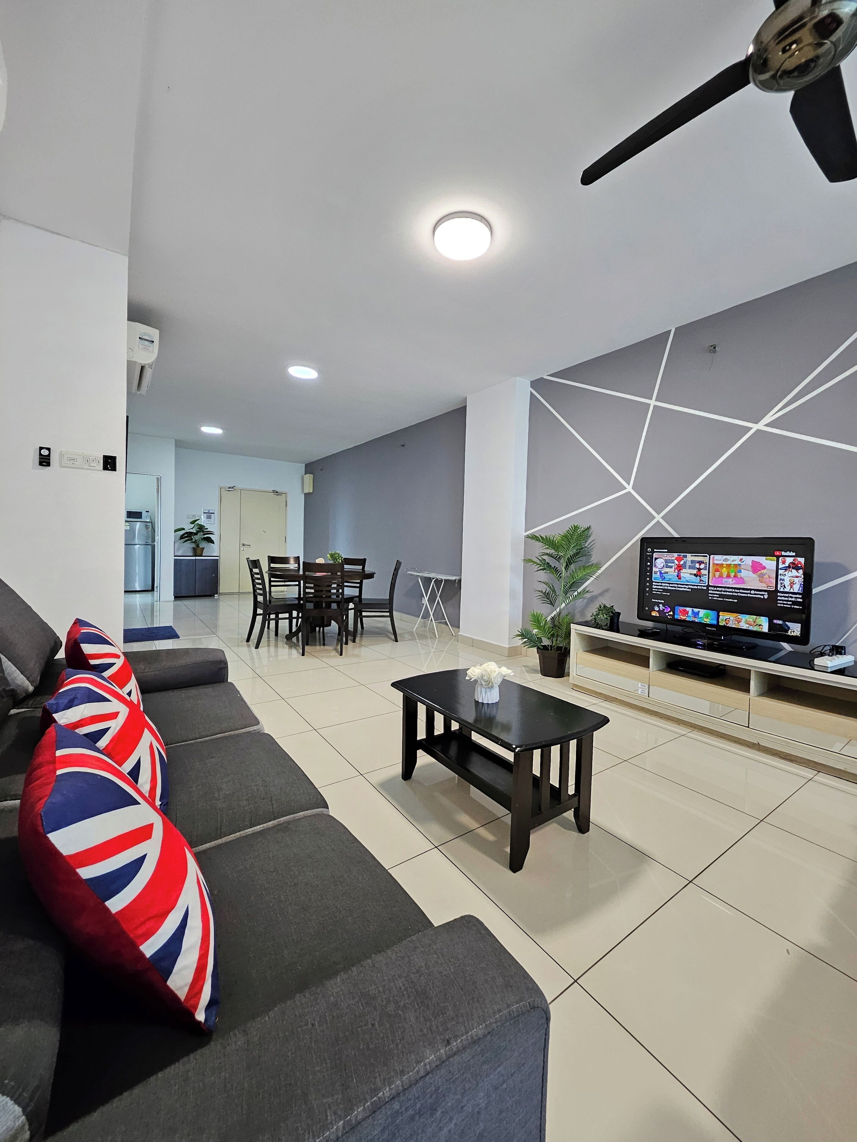 A modern living room with a grey sofa, a coffee table, and a TV on a stand. There is a dining area in the background with a table and chairs.