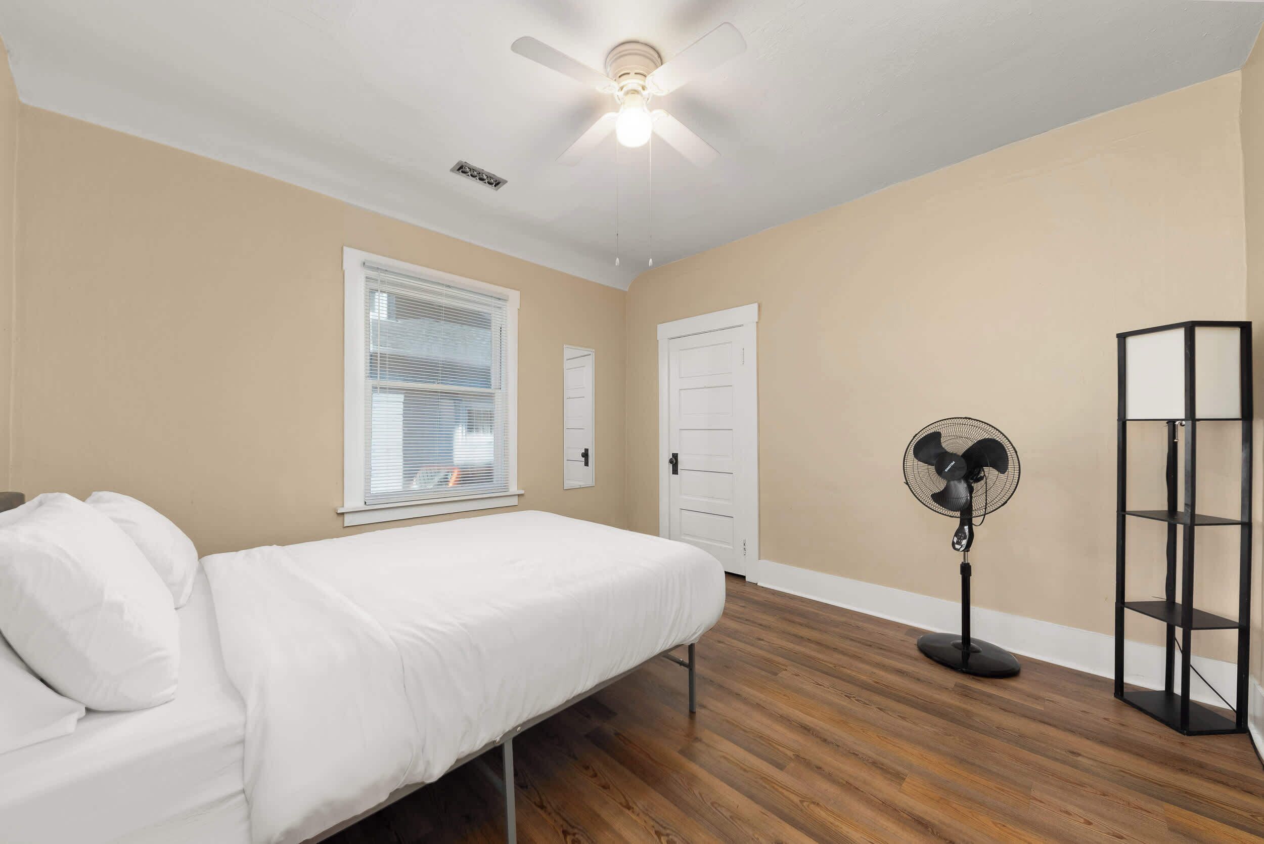 Quarto com uma cama de solteiro, um ventilador de chão, um ventilador de teto e uma janela com persianas.