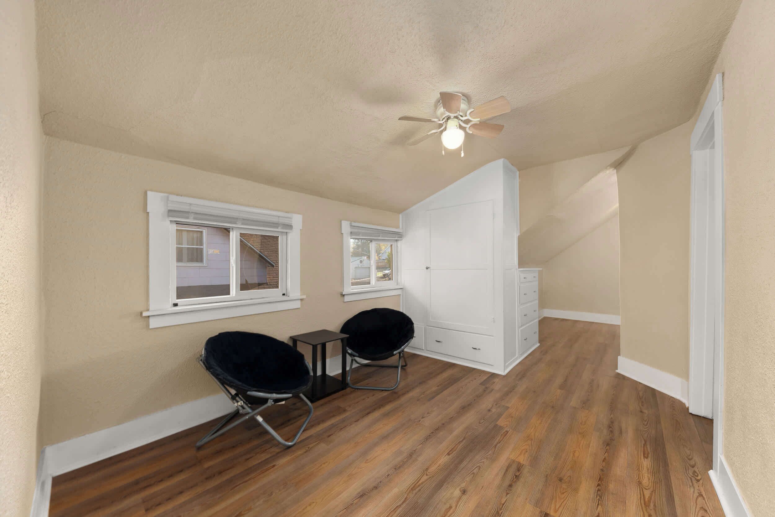 A room with wooden flooring, two black chairs, a white wardrobe, and a ceiling fan.