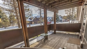A covered outdoor area with a bench, a small round table, and a view of a snowy street with houses and cars. - Cute condo w/fireplace, private decks & community pool/hot tub/sauna - Cozy Curley Bear Condo (Big Sky)