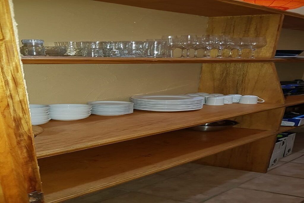 A wooden shelf with stacked white plates and cups, and a row of clear wine glasses on the upper shelf.