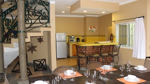 Zona pranzo con tavolo di legno apparecchiato per quattro, cucina sullo sfondo e una scala con ringhiere in metallo decorativo.