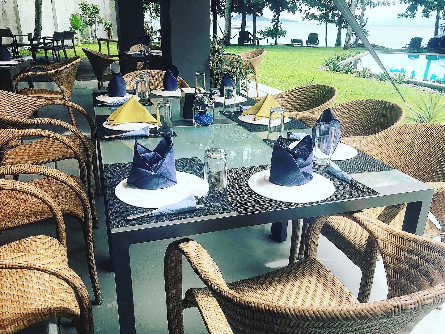 Comedor con sillas y mesas de mimbre, listo para una comida con servilletas azules y platos blancos.