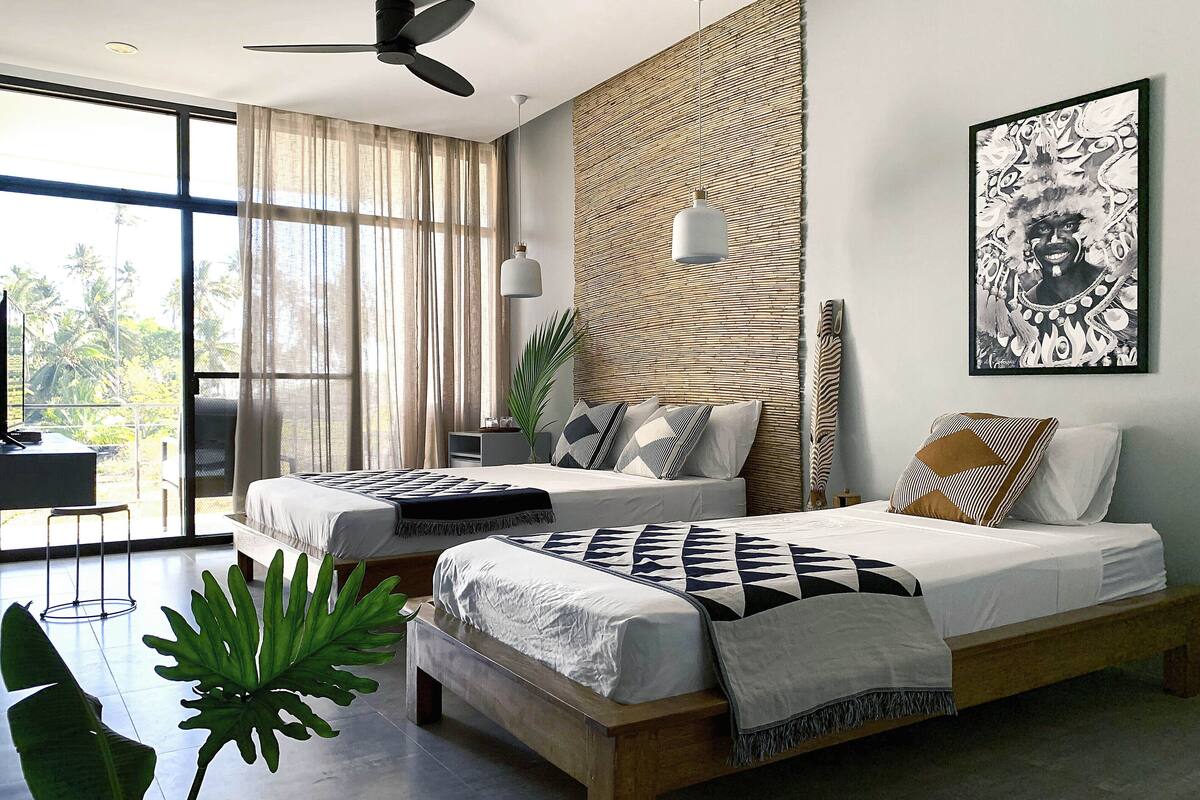 A modern bedroom with two beds, a ceiling fan, a large window with curtains, a desk, and a plant.