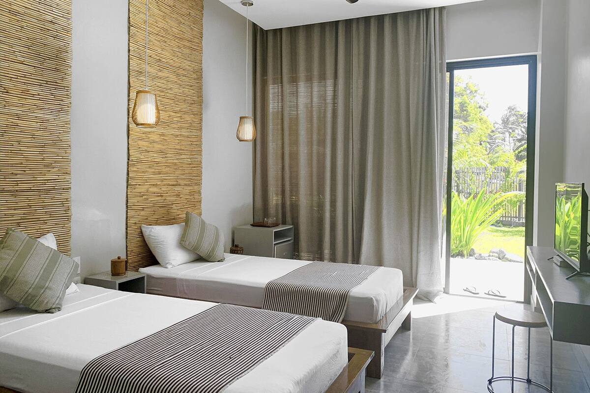A modern hotel room with two beds, a TV, and a view of greenery outside.