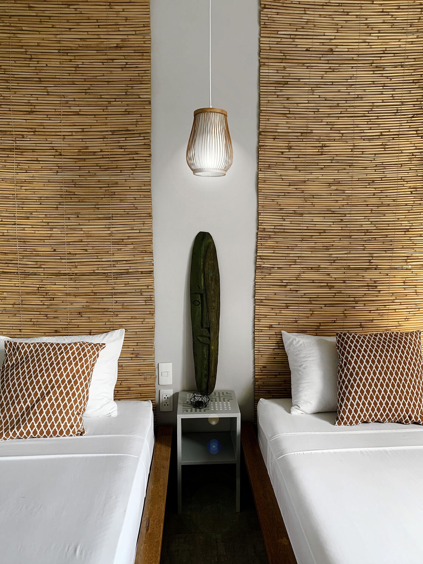 A hotel room with two beds, a wall-mounted lamp, a small table with a decorative item, and a wall with a woven pattern.