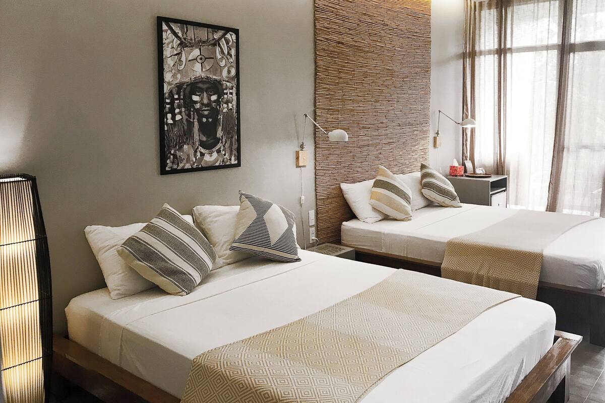 A hotel room with two beds, a large artwork on the wall, a woven headboard, and a ceiling fan.