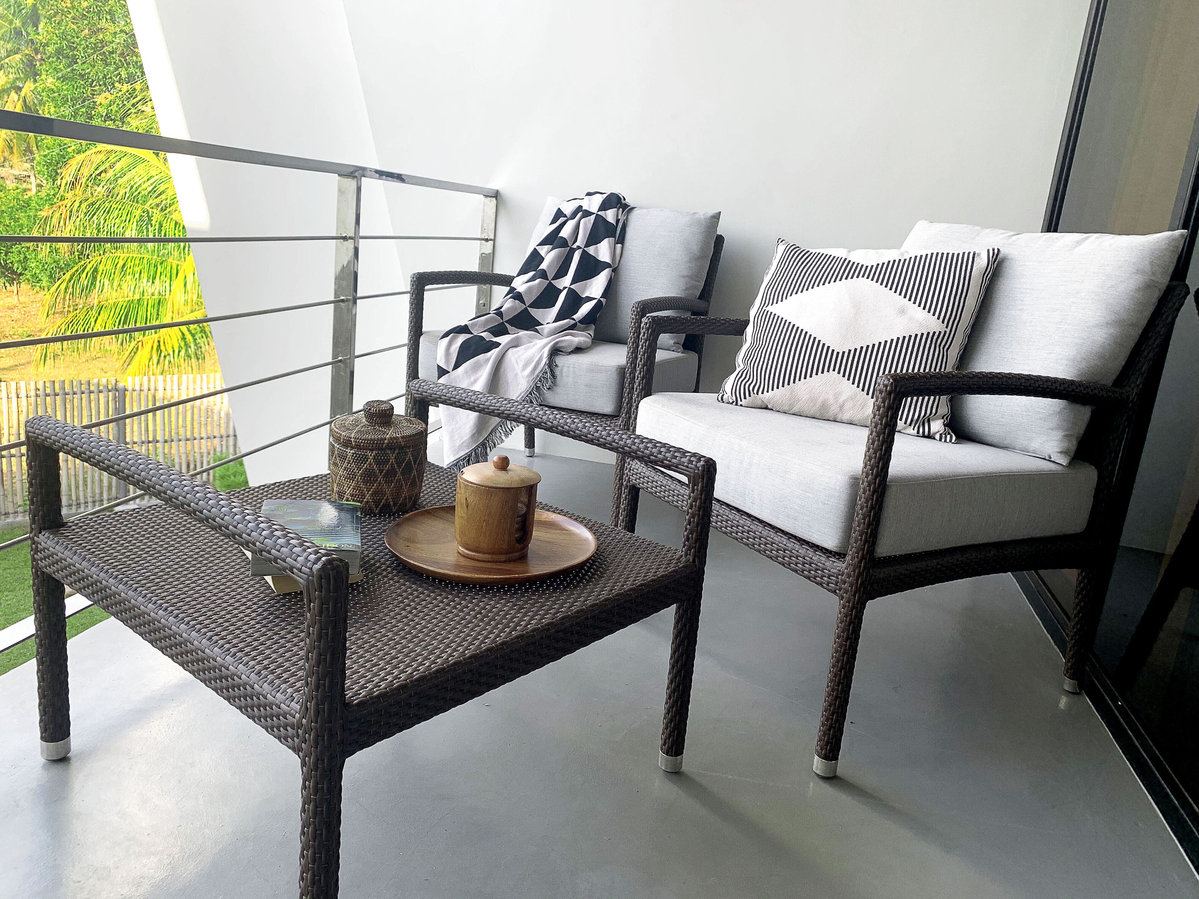 En balkon med fletmøbler, et bord med en trækasse, og en polstret stol med stribede puder.