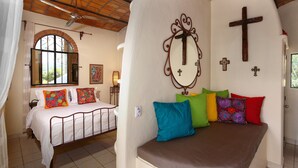 Une chambre à coucher avec un lit, un banc agrémenté de coussins colorés et un mur orné d’une croix en bois et d’un miroir.