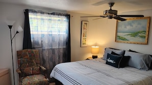 Une chambre à coucher avec un lit, une chaise, un ventilateur de plafond, une lampe et un tableau au mur.