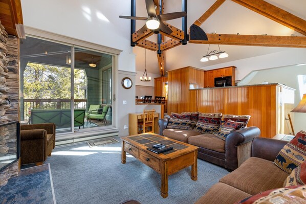 A spacious living area with a large sofa, a coffee table, and a dining table. There is a kitchen area with wooden cabinets and a ceiling fan. A large sliding glass door leads to an outdoor patio area.