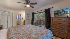 Quarto com cama, ventilador de teto, cômoda de madeira, janela com cortinas e vista para uma floresta.