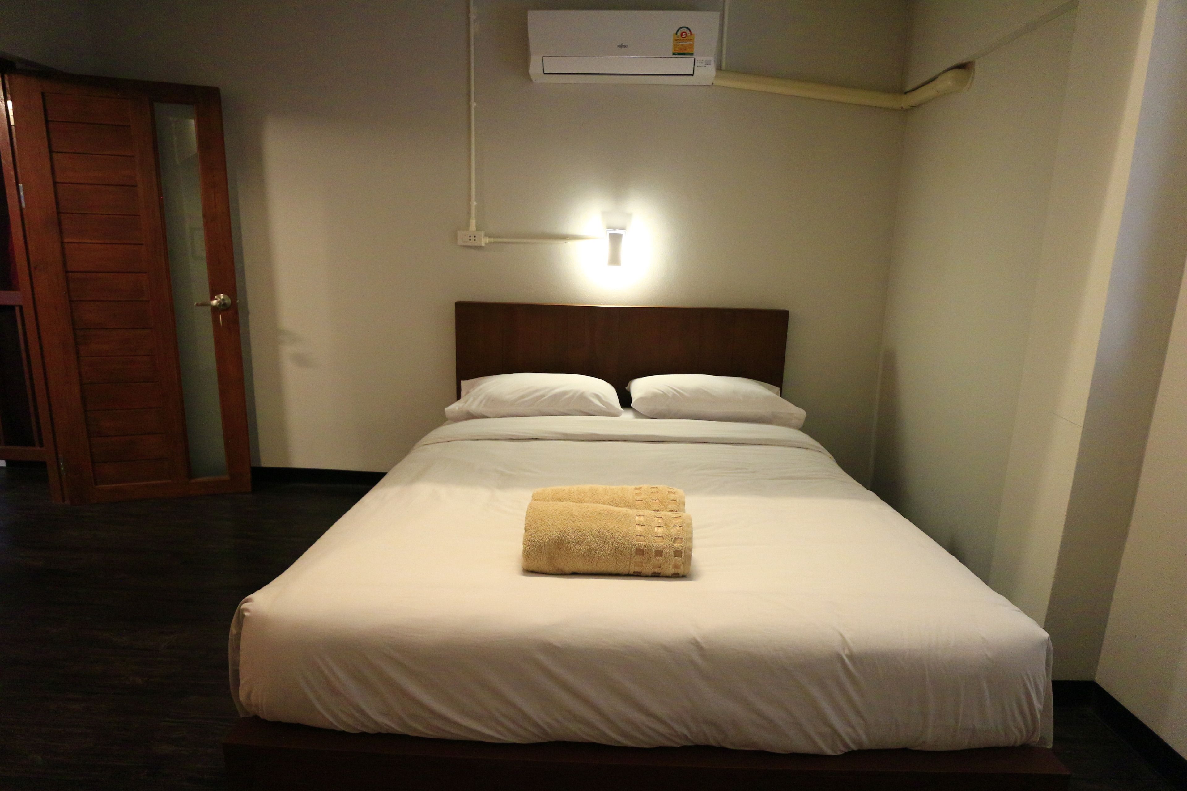 A neatly made bed with white linens and a folded towel, an air conditioning unit above, and a wooden door to the left.