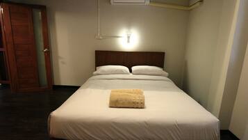 A neatly made bed with white linens and a folded towel, an air conditioning unit above, and a wooden door to the left.