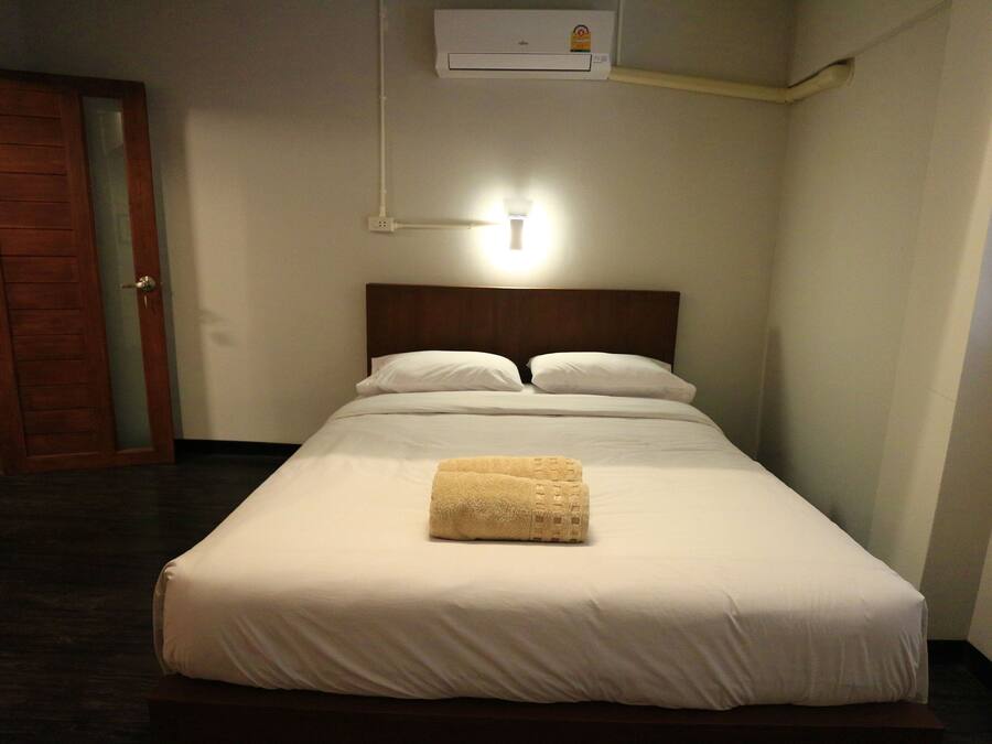 Een net opgemaakt bed met witte lakens en een opgevouwen handdoek, een airconditioning unit erboven en een houten deur links.