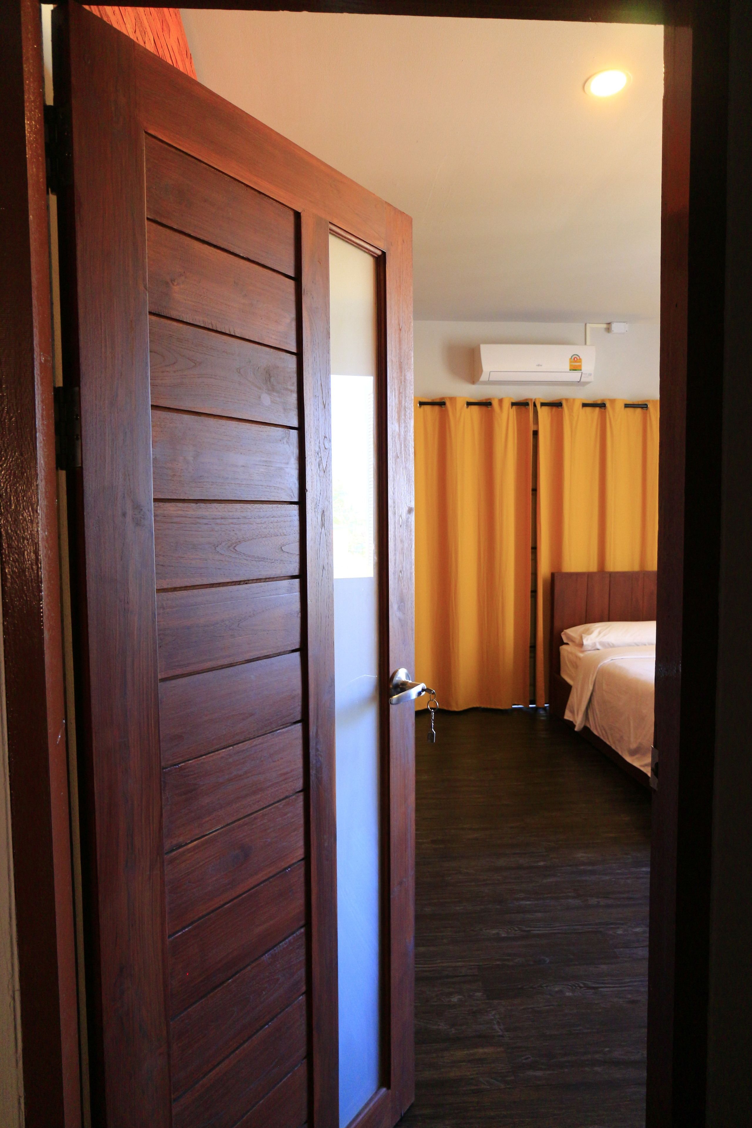 A hotel room with a wooden door, a bed, a nightstand, and an air conditioning unit.