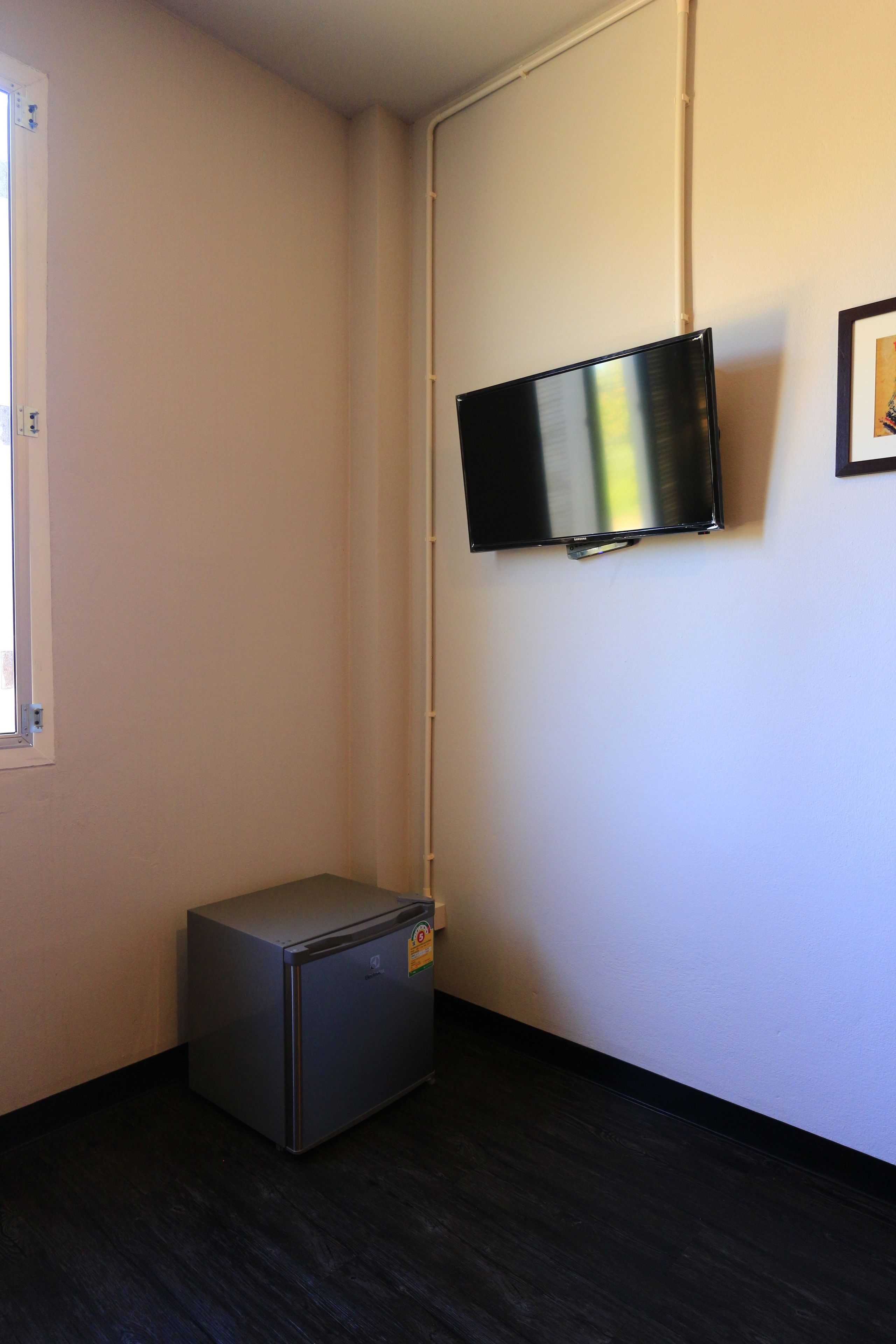 A room with a flat-screen TV mounted on the wall, a small refrigerator, and a window with a view of the outside.