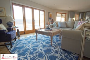 Ein geräumiges Wohnzimmer mit großem Fenster, einem blauen Teppich mit Muster, einer Couch und einem Couchtisch.