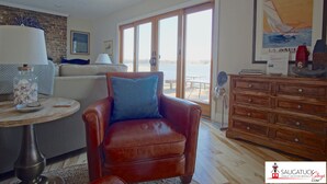Ett vardagsrum med en fåtölj i läder, ett träbord med en lampa, en soffa och en byrå i trä. Det finns ett stort fönster med utsikt över vattnet och en inramad tavla på väggen.