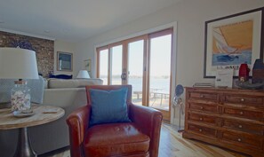 Un salon avec un fauteuil en cuir, une table en bois sur laquelle se trouve une lampe, un canapé et une commode en bois. Une grande fenêtre offre une vue sur l’eau, et un tableau encadré est accroché au mur.