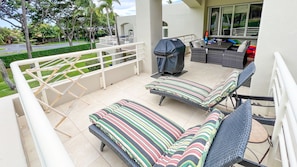 A balcony with two lounge chairs, a grill, and a view of a residential street.