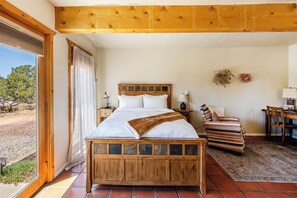 A bedroom with a wooden bed, a nightstand, a chair, a desk, and a view of the outdoors. - Stunning Desert Sunsets & Mountain Views - 19 Min, to Plaza (Santa Fe)