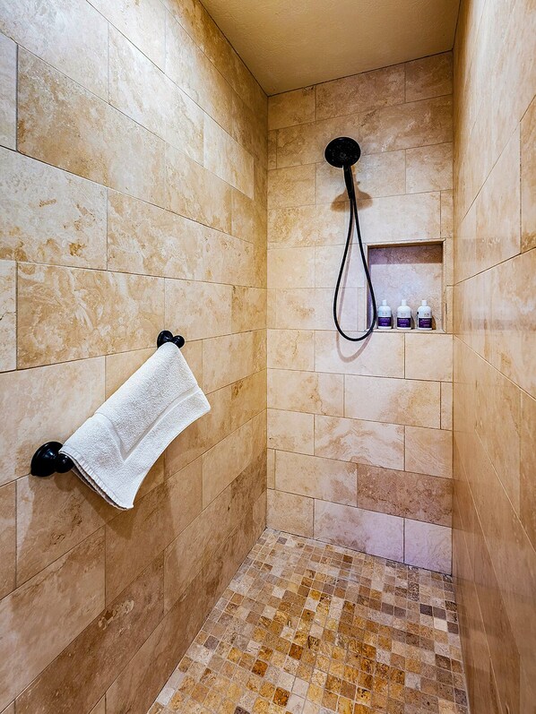 A bathroom with beige tiles, a shower, a towel rack, and a shelf with toiletries.