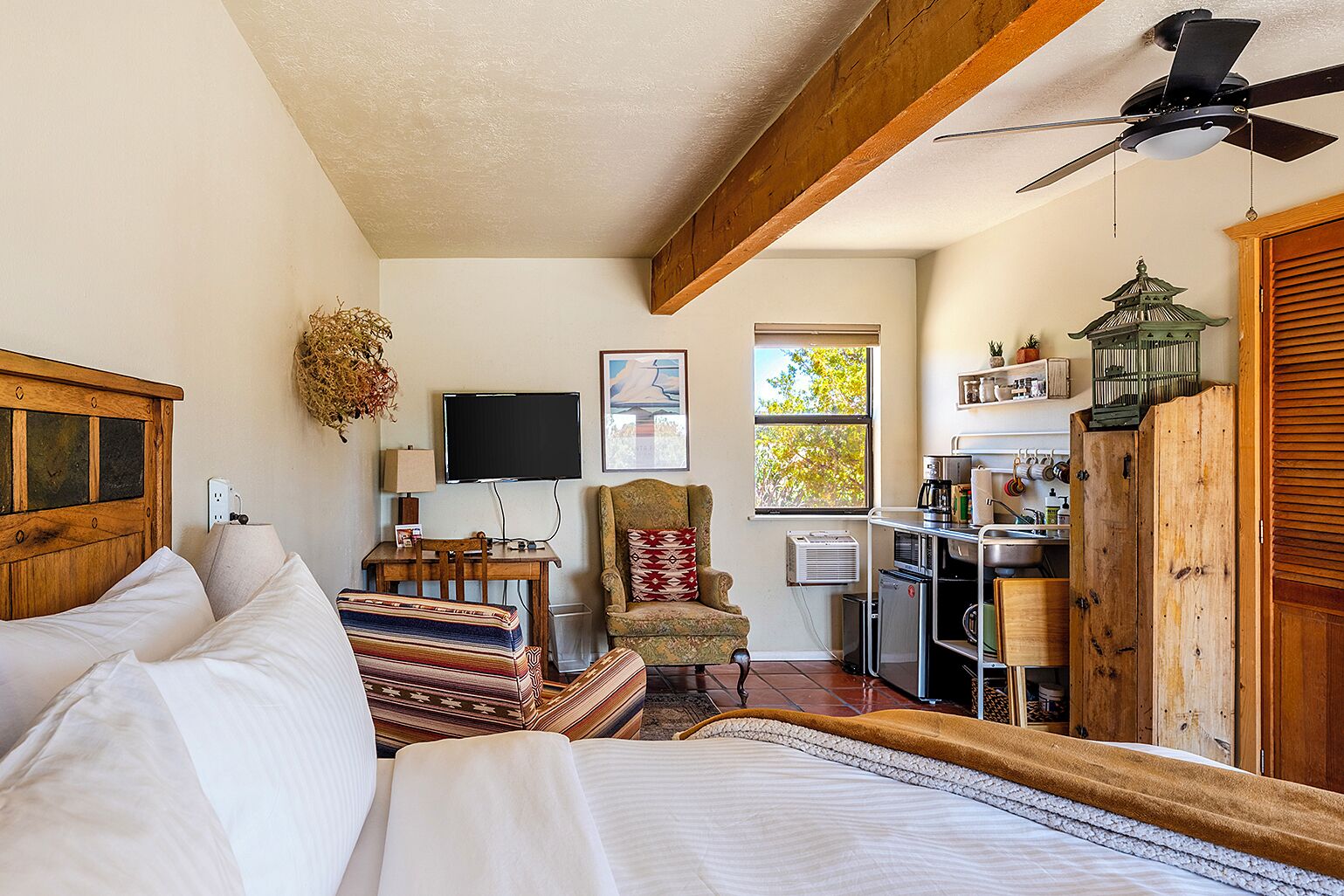 A bedroom with a bed, a chair, a desk, a television, and a kitchen area.